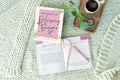 100 Days of Believing Bigger devotional journal with open pages, pink pen, coffee cup, and cozy knit blanket background