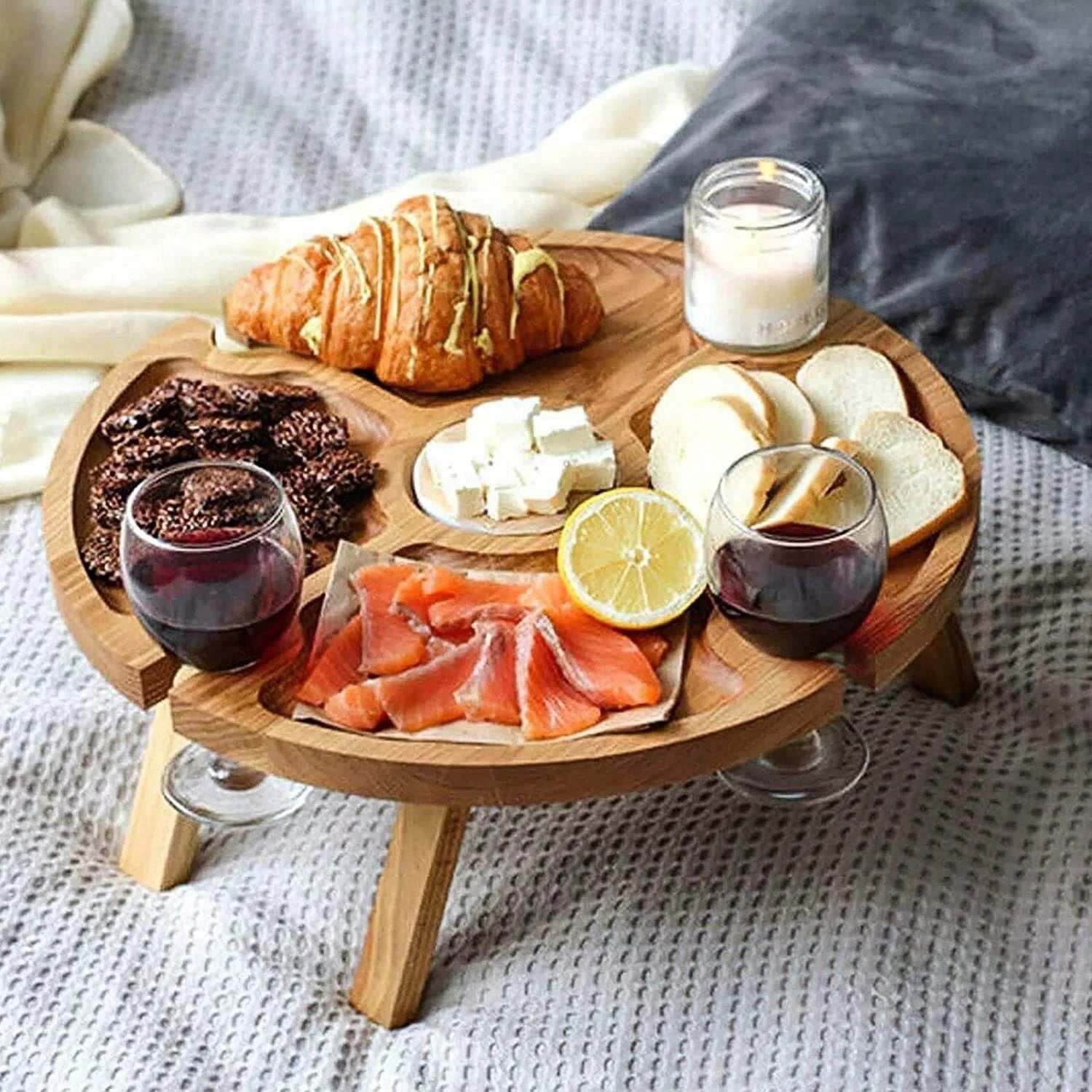 All natural collapsible charcuterie board table with glass holders serving croissants, cheese, smoked salmon, lemon, crackers, and wine